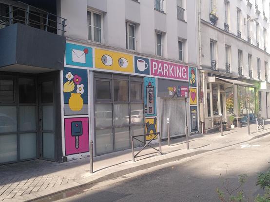 Photography of a parking entrance inside of paris. There is an automated door in light gray and a square of 9 windows, the bottom and middle ones being covered in black on the inside. Around and above are cartoonish drawing depicting a pair of eyes on a plain yellow background, a car key on a plain magenta background, an enveloppe with a notification dot on a light blue background, a black car on a yellow/orange background and a few more. A depiction of a tri-color light is made around a real tri-color light probably indicating operation of the automated door