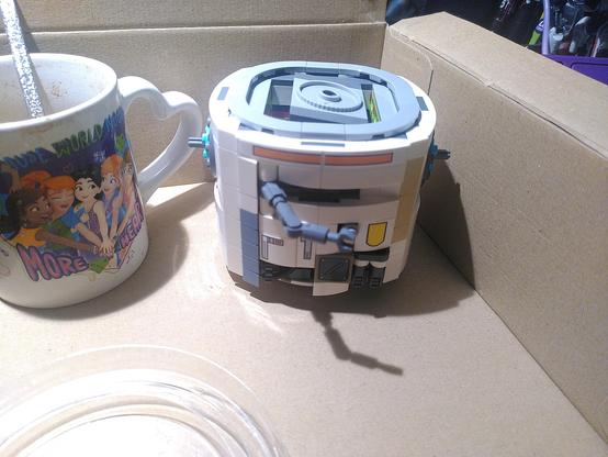 Photo of lego 75416 after bag 3/7. It's a cylinder, white with some grey, orange and yellow, a grey mechanical arm coming from the middle and on top grey circle.
It's sitting in a cardboard box. On the side is a white ceramic lego friend mug saying "because the world needs more hearts, with 5 girls, each with different skin shade, putting their hand on top if each other. The part to hold the mug is heart shaped.