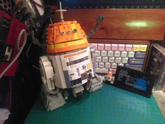 Photo of lego 75416 finished. It's chopper, the astromech from star wars rebels. It have a short white torso, two legs and a central wheel, an orange head with two arms, raised in protest in the photo and an antenna.
On the side is a plaque with some info on the droid and a minifigurine version. In the background is an unrelated mechanical keyboard with a wood casing and palm rest