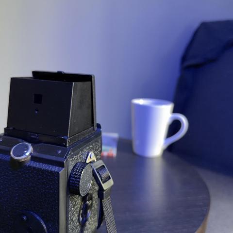 An old TLR film camera sits patiently waiting for use whilst the subject is a white mug and a chair on which someone has draped a coat.
