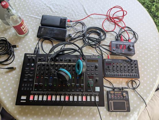 A table full of music gear. One big device full of colorful LED pads, fader and pots is my Roland Tr-8S drumcomputer. Right next to it are three devices, on top a midi clock set to 133 beats per minute. Below it a small synthesizer, a Roland Aira compact T-8 which kinda looks like the Tr-8S but without the faders and waaaay less potis and LED colors. It's also tiny compared to its big brother. And below that comes a Korg NTS3 Kaoss Pad which looks like a laptop touchpad with a few extra buttons and a LED display.