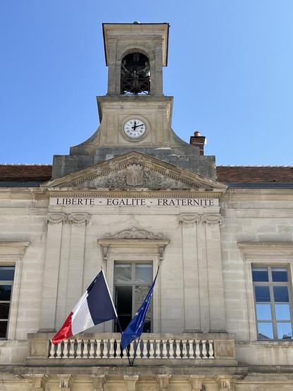 Le haut d'une mairie dans un bâtiment ancien avec horloge, clocher et bien sur Liberté Egalité Fraternité gravées, et drapeaux Fr et Eu.