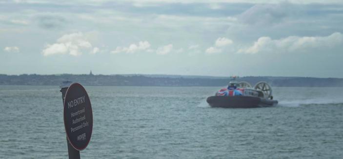 An image of a hovercaft approaching a beach hoverport.  The hovercraft has a black skirt and and predominately white body with a union flag painted on the front.  In the foreground of the image is a sign stating "No entry.  Hovertravel Authorised Personnel Only"