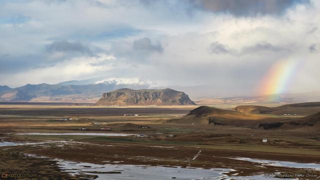 A colour photo of a view from height overlooking a large flat area of land with some prominences. The sky is overcast but strong sunlight is coming from the left, and the landscape is dotted with cloud shadows. In the distance is a glacier-capped volcano, only partly seen due to heavy cloud at its cap. The foreground is brown meadow land with large areas of ice and snow, in one place a farm road is highlighted in ice. Several buildings are scattered around, mainly small clusters of farm structures. In the middle distance is a slope-sided brown rock island with vertical surfaces near the top. To its right is an arc of strong rainbow colours, hanging above another prominence.
