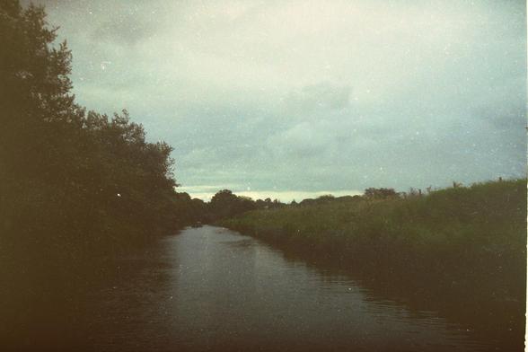 This is a faded image of a photograph circa 2003.  It is taken from a canal boat moving slowly down the kennet and avon canal in wiltshire.  The left hand side is a tree lined bank, the right hand side is farm land.  The sky is over cast and there is no sign of any sun.  The image, which is scanned from a negative is faded and a bit scratched.