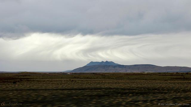 A colour photo of a landscape under heavy overcast. Thick grey clouds are swirling overhead, a clearer space below them. In the distance is a small mountain and the foreground is brown vegetation and black gravel. In the gap between the cloudbank and the ground surface is an odd, almost right-angled stream of cloud, probably rainfall caught in the wind and skewed by it. At the top it's falling towards the left, then makes a sharp dogleg turn towards the right. It's quite surreal.