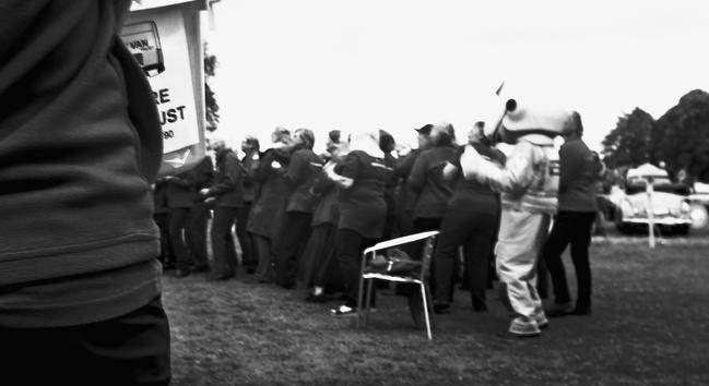 A community choir in full voice sings out in the background. Next to them stands someone in a bear costume.  The bear is wearing a flight suit and flying helmet.  In the foreground is a volunteer for a charity holding a collection bucket.  The image was made at a charity event in aid of the Wiltshire Bobby Van Trust and the Wiltshire and Bath Air Ambulance.