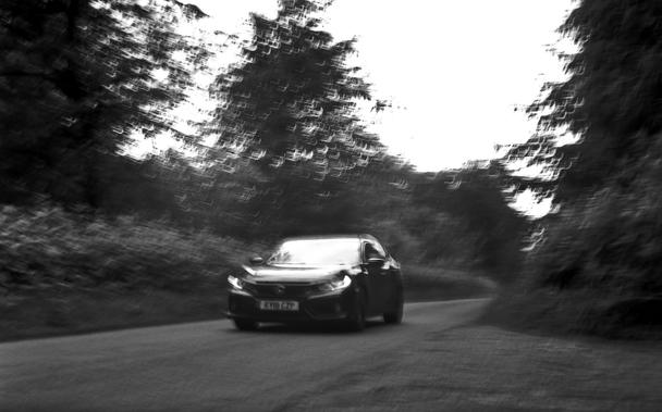 A black and white image a car racing up a hill there are trees on either side that are moving as is the car.  The whole image is an example of extrusive time