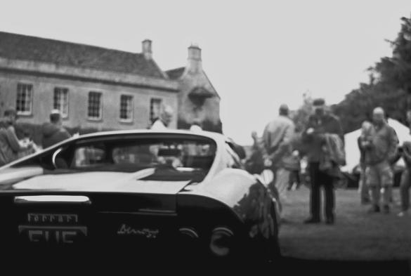 The scene is a of the rear of a Ferrari Dino which has been parked on someone's lawn.  There is a posh house in the background and there are lots of people looking at the car.  Only the car itself is in focus the rest of the people are just blurs.