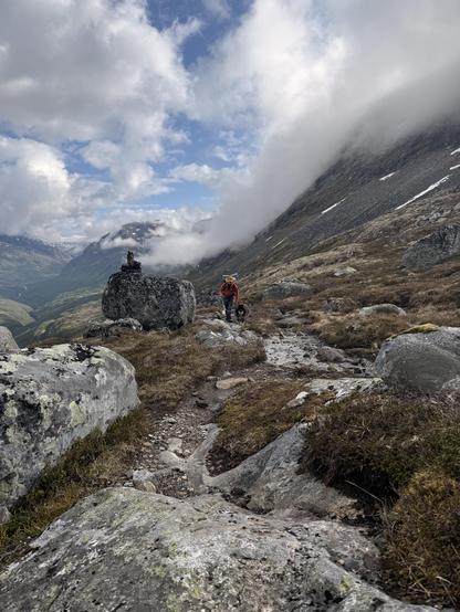 A man with hiking gear and a fluffy black dog climbing up a rocky path surrounded by mountains.