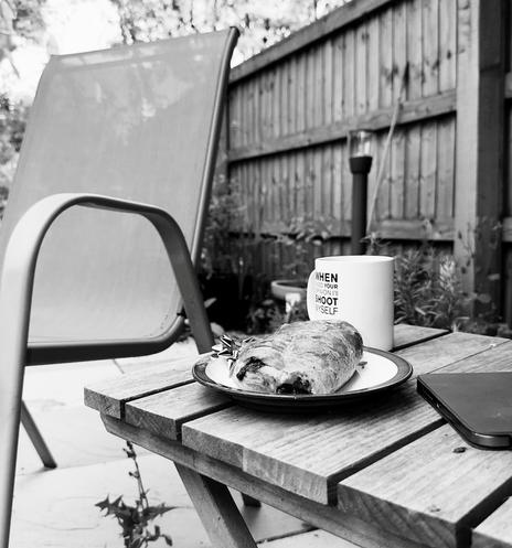 A garden chair empty of its occupant.  Next to it is a small wooden table on which is a pan au chocolate, a mug of coffee and an iPad.