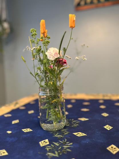 A small flower arrangement of California poppies (orange, petals closed), tiny white cilantro blossoms, green oregano blossoms, and pink and white clusters of another wildflower whose name escapes me. The vase is an upcyclyed peanut butter jar.