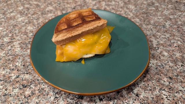 Picture of a granite countertop and a small green plate with a light brown trim. On the plate is a sandwich made out of two quarters is a round waffle. There's a yellow cheese melting over a fried egg in the sandwich.