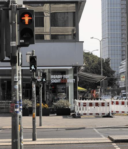 An early morning crossing in Berlin showing the two forms of the Ampelmännchen - the pedestrian crossing symbols. In the foreground is the stop - a red figure in a hat has his arms out stretched, T-posing  if you will. In the background is the cross form. The figure is walking now, caught in mid stride and with one arm out. The other arm though isn’t posed.