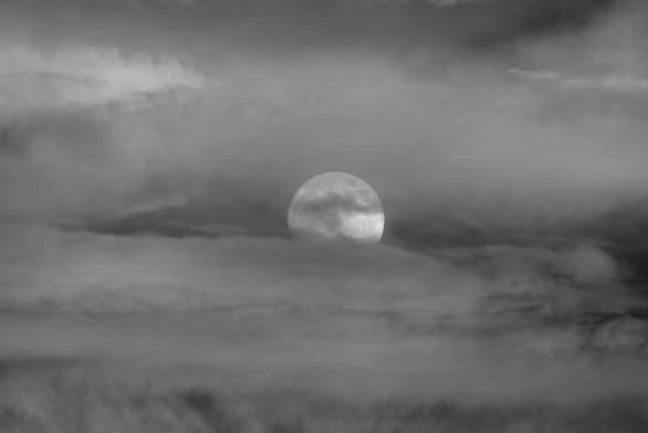 The full moon rises through a break in a cloud bank.

This is a daytime shot. The sun was barely above the horizon and illuminating the distant clouds.

A small ragged cloud covers part of the moon's face, and the bottom third of the moon is hidden by a ribbon of cloud.