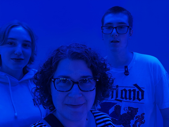 Me and two kids in blue light (white lady with glasses and curly hair between tall white teen boy in a Scotland shirt, buzz cut and glasses, and white blond girl in a hoodie