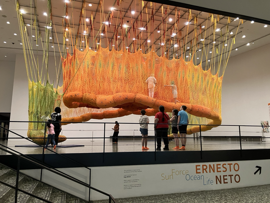 Art thing; orange lumpy tube spiraled around the room and hanging from the ceiling by a bunch of cord thingies. People are climbing in it. It’s like a woven or macrame wall on each side and a tube of ball pit balls at the base. Very organic and very swingy inside and fun and cool