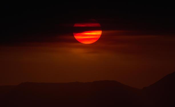 An orange-red sun is half covered by distant thick smoke clouds.  The foreground mountain ridge below the sun is a dark silhouette. Orange-red is the only color in this image.