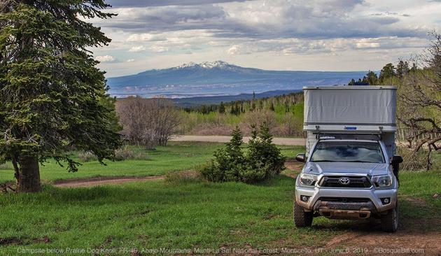 A pop-up camper with lifted roof sits on a small, silver pickup truck with nose toward the camera, on the right side of the photo. A large evergreen tree with a thick trunk stands on the left. Beyond this grassy area is a sparse woods and beyond that a broad conical mountain with a touch of snow on the peak. Heavy clouds.
©BosqueBill.com