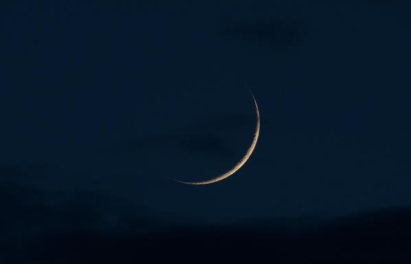 The cresent moon is yellowish because it's not high above the horizon, and it's a bit smoky from forest fires in Utah. The moon sits in a dark blue twilight sky with some small clouds near it and a layer of darker clouds below it.
