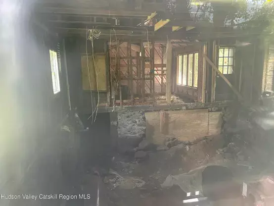 Photo, clearly taken through a window, of a large interior of a home with no dividing walls, no interior surface on the studs for the exterior walls, no floor or even joists above the basement. The basement has very rough walls like a concrete foundation just made in a ditch and an uneven dirt floor with large rocks strewn about. It looks like someone tried to shore up the basement wall with plywood
