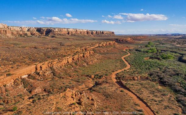 A dirt road winds along a valley roughly parallel with a dry wash. The only side of the valley in the aerial photo is comprised of red sandstone cliffs, a sloping terrace, then white sandstone cliffs. There are desert shrubs and small trees on the valley floor near the wash. Blue sky with a few clouds.
©BosqueBill.com