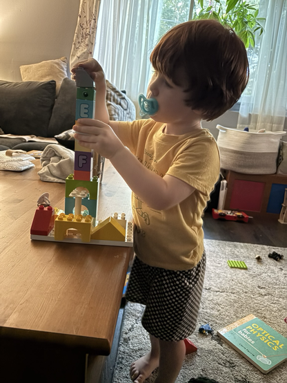 A little boy in a yellow shirt playing with Duplo blocks. He is making a tower that is as tall as he is