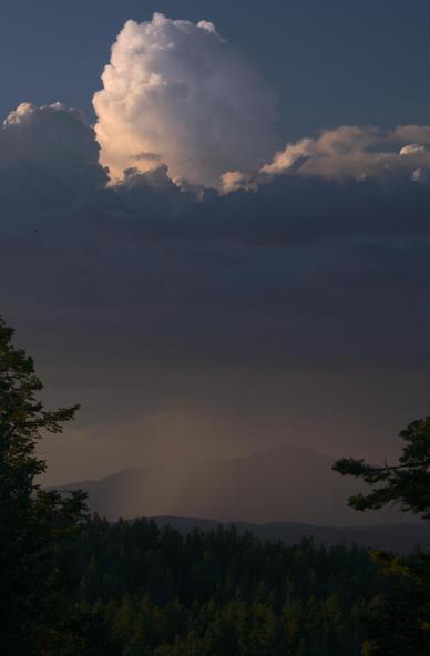 A monsoon thundershower stands tall and catches the last sunlight of the day as shafts of rain, muted and not as bright as the cloud tops, fall on distant rolling terrain. Sierra Blanca peak is a dark silhouette behind the rain.