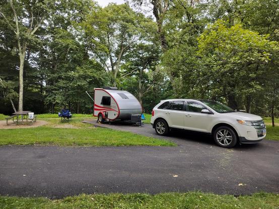 Small red and white camper behind a white SUV on a campsite surrounded by trees