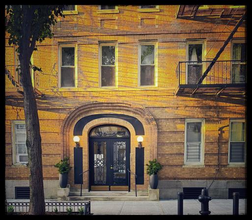A color photo showing the front of a building with a rounded arch of a doorway, but looking a little bit modern. The arch is made of stone but there is a black metal strip in the middle with a round globe light on each side. The doors are black iron with a design. The sun is hitting the yellow building casting shadows from fire escapes that are on the left over horizontally to the right. A bit of a fire escape can be seen on the right and on the left side is a single tree with a bit of green leaves at the top.
