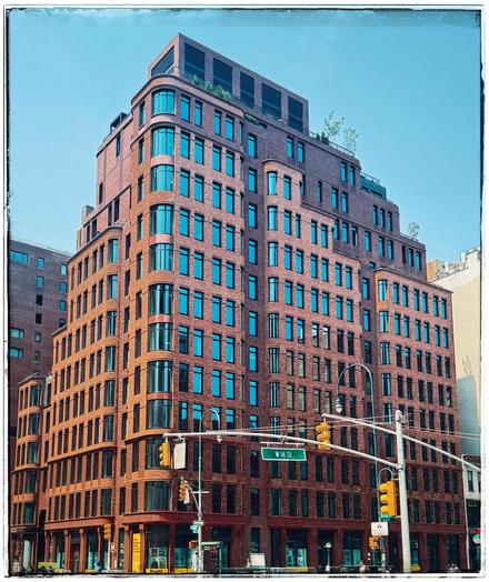 A color photo of a large brick corner building. It is 12 stories high, with a smaller square structure on the top. It is all a reddish orange brick with vertical rectangular windows spaced a few feet apart. On the corner the windows and the brick are all rounded. it almost looks like it could've been here for a very long time, but is brand new. There is a lamp post towards the bottom and also a stoplight hanging over the middle with a green sign that says 14th St. On the roof of the building some greenery can be seen sticking out just a little bit and there is a clear blue sky above.