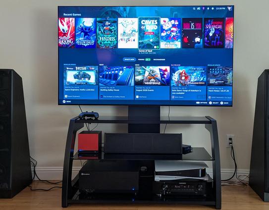 Picture of the Steam UI running on a large oled TV, with an orange-faceplated Steam Machine below it, sharing space with a center channel speaker, HTPC box, Playstation 5, and A/V receiver.