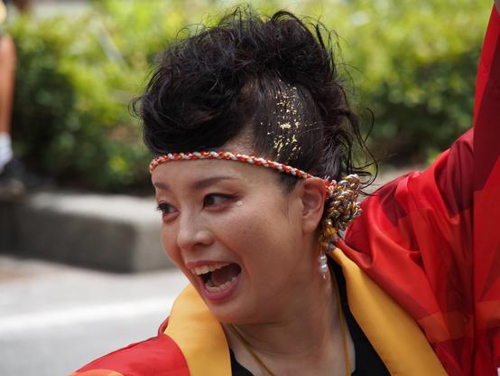 Close up of a festival dancer in bright red and yellow costume. She's looking to the left, arms raised, shouting along to the music.  Her hair is flecked with gold leaf, and tied with a white, red, gold and silver cord.