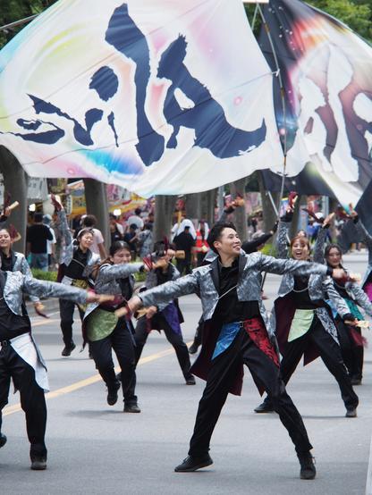 A dozen festival dancers performing on the street, with very large festival flags waving above them, one white and one black, each with kanji surrounded with rainbow colours. The dancers wear modern styled costumes, black with shiny silver jackets, and coloured accents around their waists. Their arms are flung out mid-dance, and the most prominent dancer in the foreground is smiling widely and looking off to the right.