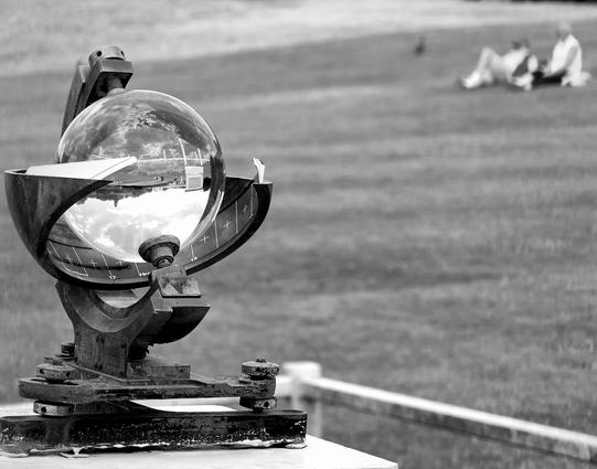 A brass and glass Campbell–Stokes heliograph (sunshine recorder) sits on a white plinth in the foreground. In the background (and out of focus) are two people having a picnic and being observed by a duck.  

The recorder consists  of large ball lens mounted into a metal housing that allows concentrated sunlight to be focused on a strip of card that is then charred by the sun.  It is designed to record how many hours of bright sunshine occur on a given day.