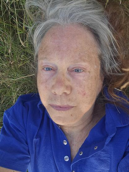 Portait of an old woman, pale with blue eyes, blue shirt, grey hair lying on her back in a grassy sheep pasture listening to music.