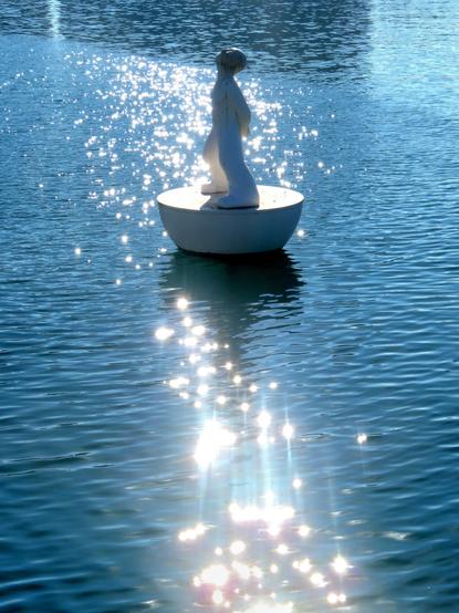 The image shows, from a slightly higher angle, the sculpture "Miraestels" ( Stargazer ), a work by the artist Robert Llimós. It consists of a hemisphere with a figure of a child gazing at the sky resting on its flat side. It is made of white plastic resin. The sculpture is designed as a floating buoy in the port of Barcelona and is a tribute to the world of the Poet Joan Brossa. The photograph is backlit, and the sun produces countless flashes of light on the water.