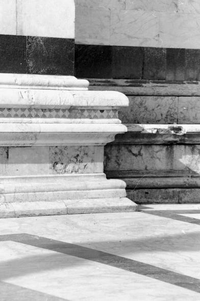 The "analog" black and white vertical format photograph shows a partial and very close view of the lower part of the exterior side wall of Siena Cathedral with a beautiful combination of the very diverse types of stone used in its construction as well as part of the floor mosaic.