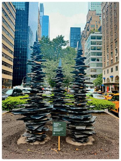 A color photo of an art exhibit on the median in between Park Avenue. There is a small green sign in front that says Manolo Paz 2025, but the small wording underneath is unreadable. There are three 6–7 feet tall stacked rock formations. It is all done within pieces of black slate, not larger round stones. The formations start out medium and get a little bit bigger and then smaller as they go up to the top. They are sitting in brown sand that has been rounded out a bit for them to sit in the middle. Behind them across the middle is some greenery And a large green tree stands behind them. There's much traffic going up and down on either side of the median. Many tall buildings can be seen receding into the background with a very light blue sky at the top.
