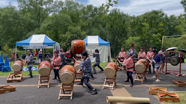 A dozen taiko performers, young and old, in traditional red and blue Japanese festive outfits play various taiko drums: a large one rear center, six on stands in front, and four smaller ones on stands in a row off to the right. They are set up on a broad sunny path along a grassy park area with trees and some blue and white festival tents in the background.