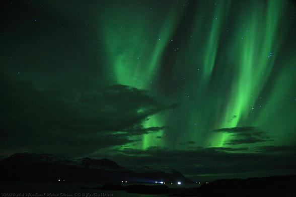 A colour photo of a night sky with lots of stars. At the bottom of the shot is a group of lights from farmhouses and other buildings along the shore of the fjord. There's quite a bit of cloud above them but most of the sky is clear, and strong bands and waves of green aurora are streaking upwards from them and passing overhead. It's very lovely.