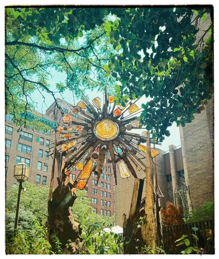 A color photo of a large sun made out of welded iron pieces and fitted yellow and orange pieces of glass. In the center of the sculpture is a large round yellow piece of glass attached with some wire to an outside piece of metal. Protruding from that are many individual metal spikes. The different length spikes have different shaped orange and yellow pieces of glass wedged in between them and attached by wire. It is being held up by two pieces of metal on either side that almost look like chunks of rock. To the right side is a little bit of the light brickwork from the front of the church. On the left side is a street lamp with a basic brick building behind it. There's lots of greenery hanging over the top of the photo with various leaves from different types of trees. A little blue sky is poking through.