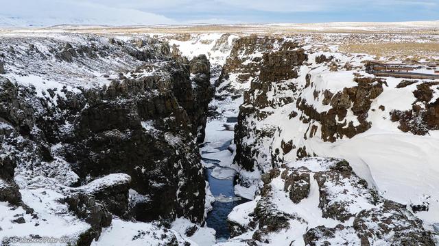 A second colour photo of the canyon looking in the opposite direction. The gorge has dramatically steep walls, quite vertical in places. The small amount of sky that's visible is grey and overcast with snow cloud. On the right is a viewing platform with a barrier and seating which provides some scale. Every ledge that's even close to horizontal is covered with deep snow drifts, but the dark rock of the canyon walls is relatively clear of ice. At the bottom of the gorge, most of the river is frozen over except for small areas where the water surface can be glimpsed.