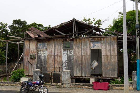 Das Bild zeigt ein zwerfallenes Holzhaus mit verrosteter Wellblechverkleidung. Das Dach ist zum großen Teil eingestürzt. In die Vorderfront sind drei große Flügeltüren mit Oberlicht eingelassen. Die rechte ist herausgerissen. 
Vor dem Grundstück steht noch ein eisernes Tor, das von zwei Betonpfeilern gehalten wird. Jeder Torflügel besitzt eine Eisenplatte. Auf dem linken ist der Buchstabe "F" zu erkennen, auf dem rechten ein "M". Der restliche Zaun fehlt.
An der Straße vor dem Grundstück wurde ein Moped mit Helm abgestellt.