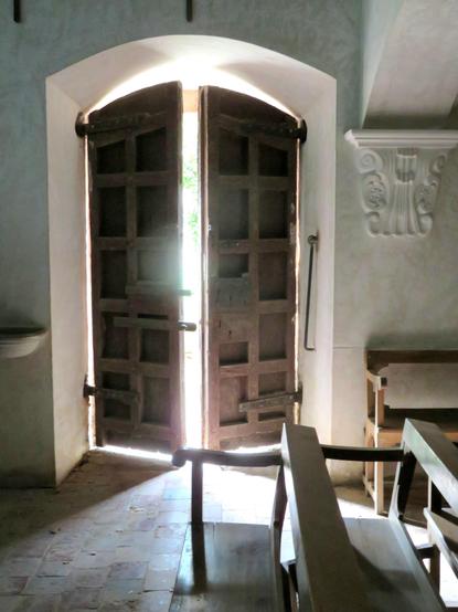 The photograph shows, from inside a dark hermitage in Vall de Bianya with white walls and wooden benches, the large entrance Door Ajar, allowing sunlight to enter and illuminating the room with a play of light, shadows and dimness.