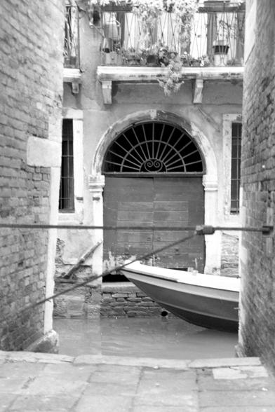 The black and white, vertical-format "analog" photograph was taken from a narrow, brick-built access point to a small Venetian canal. Through it, one can partially see the water, a gondola, and a building in a frontal view. Windows and a balcony with flowers are partially visible on the building. In the center is a large rectangular entrance door with a semicircular grille above the arch.