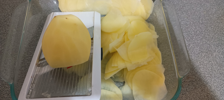 A glass dish with thinly sliced potatoes on the right and top of frame, and on the left an OXO mandoline with a potato on it