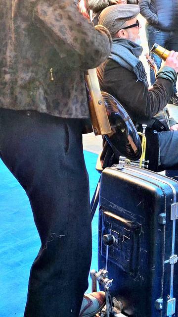 The photograph shows a detail of a Street Band seen from behind, in which a musician complements the washboard action with a pedal bass drum built by recycling an old suitcase.