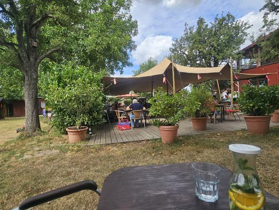Blick auf eine Wiese mit Büschen und Bäumen, im Hintergrund ein Holzhäuschen auf einem Spielplatz. Im Zentrum ist über einer großen Holzterasse eine Zeltplane aufgespannt, unter der an zusammengewürfelten Mobiliar Menschen sitzen und essen, trinken und quatschen. 
Im Vordergrund ist – etwas außerhalb – ein Teil des Tischs zu sehen, an dem ich sitze. Es steht eine kleine Karaffe mit Wasser darauf, in dem eine Orangescheibe und frische Minze schwimmen, daneben ein Glas.