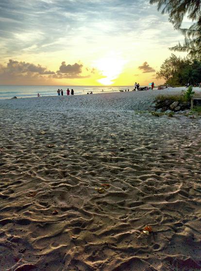 Sunset on a beach in Barbados. Small clouds on the horizon and, far off on the beach by the water, the silhouettes of several people. To the right, local vegetation. The majority of the photograph is taken up by the beach sand, which, backlit by the setting sun, creates a beautiful visual texture.